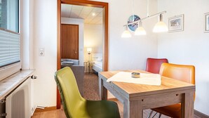 Een eetgedeelte met een houten tafel, stoelen en hanglampen. Een raam met jaloezieën is links zichtbaar.