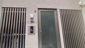 A modern building entrance with a textured facade, a metal door with a security keypad, and vertical blinds on either side. - New open F Maison Kishinosato 301 (Osaka)