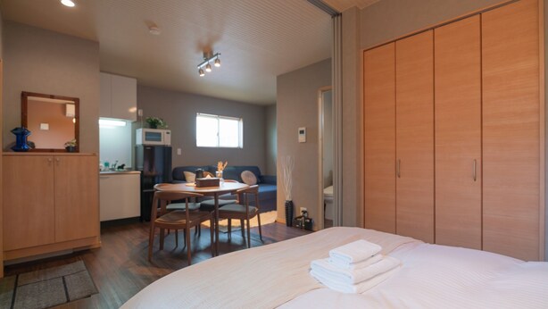 A modern bedroom with a large bed, wooden wardrobe, and a small dining area with a table and chairs. - New open F Maison Kishinosato 301 (Osaka)