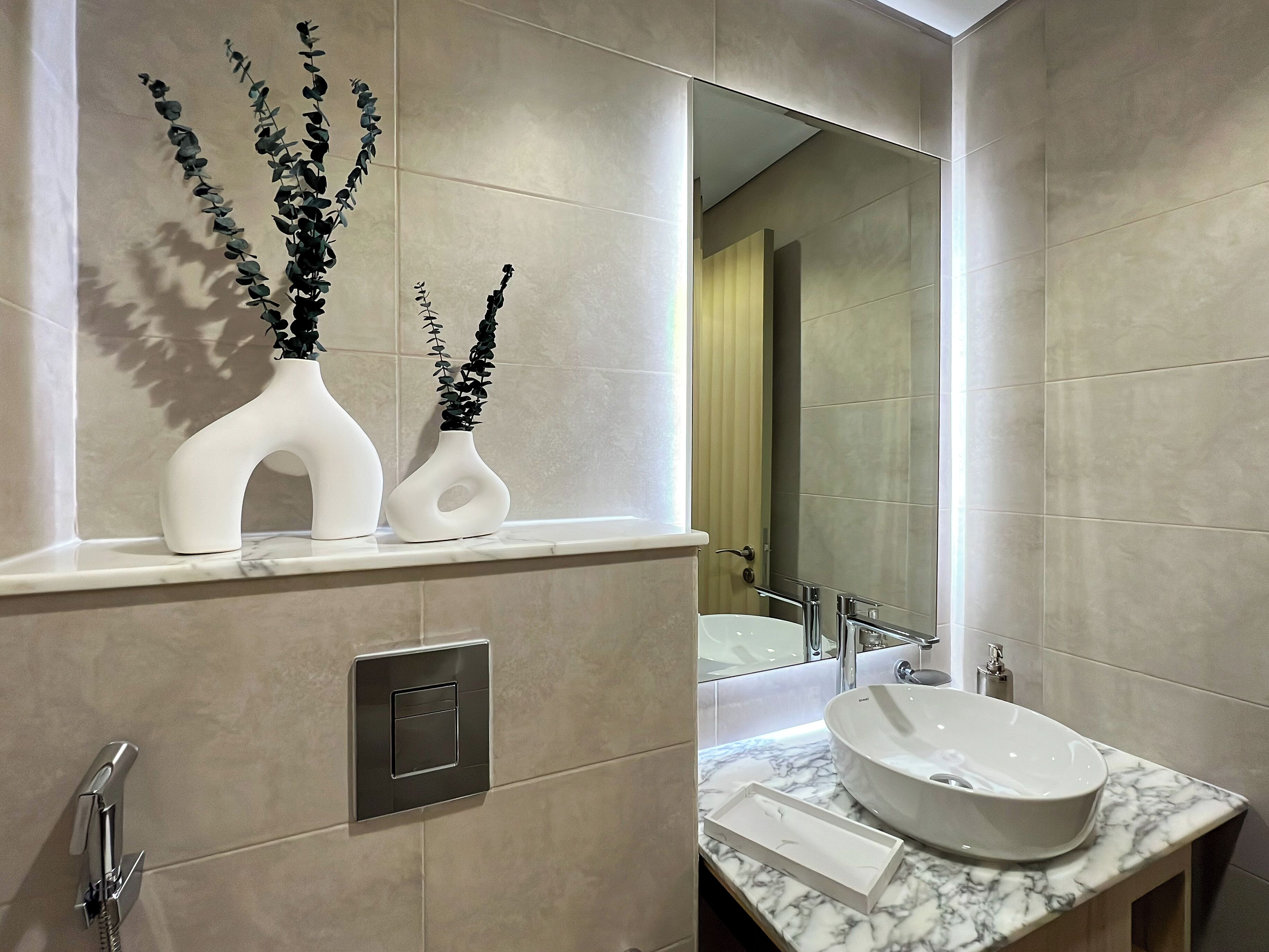 A modern bathroom with a large mirror, a white sink, and two white vases with black foliage.