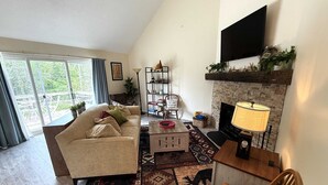 A living room with a sofa, a coffee table, a fireplace, a TV mounted on the wall, and a large window with curtains. - 2 BR Jiminy Peak w Mountain View Sleeps 7 Gorgeous (Hancock)
