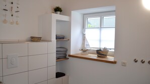 A bathroom with white tiles, a window with blinds, a shelf with towels, and a hanging wind chime.