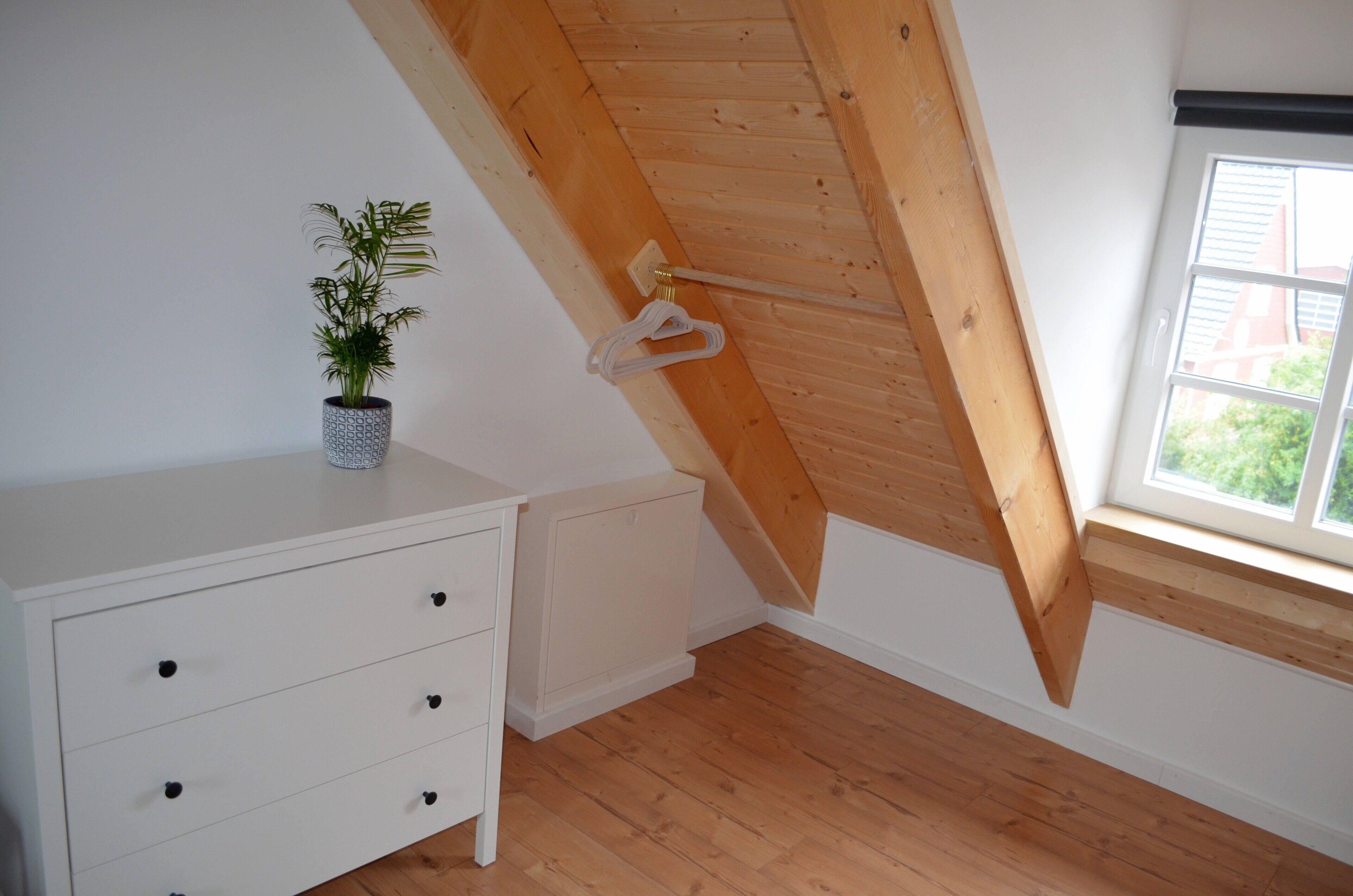 A room with a sloped ceiling, a white dresser, a potted plant, and a window with a view.