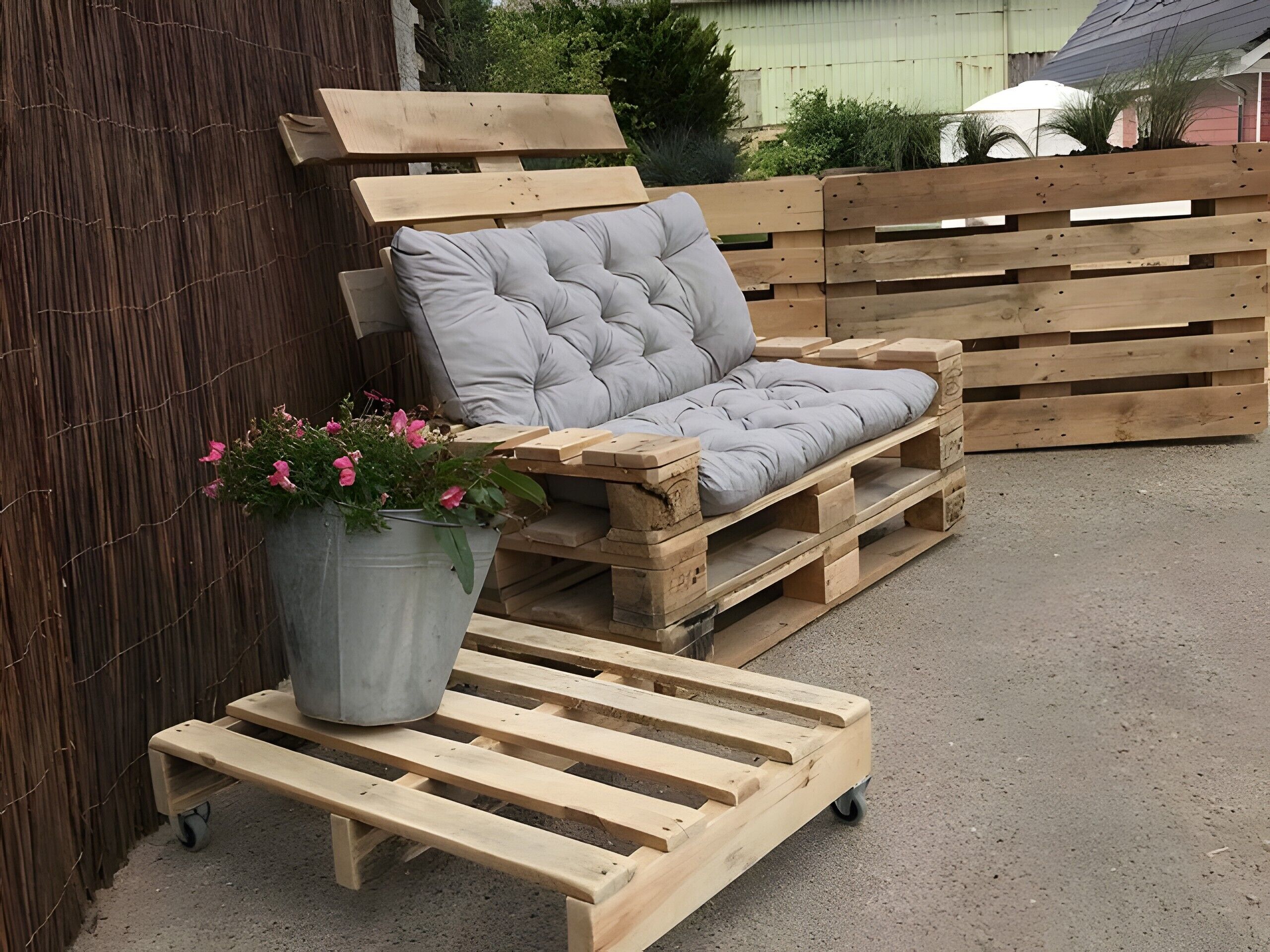 A wooden pallet sofa with a cushion, a metal pot with flowers, and a wooden pallet table with wheels.