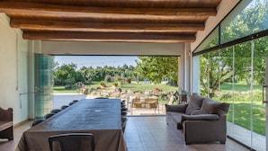 Ein offener Wohnbereich mit Esstisch, Sofa und großen Fenstern, die einen Blick auf einen Garten bieten.