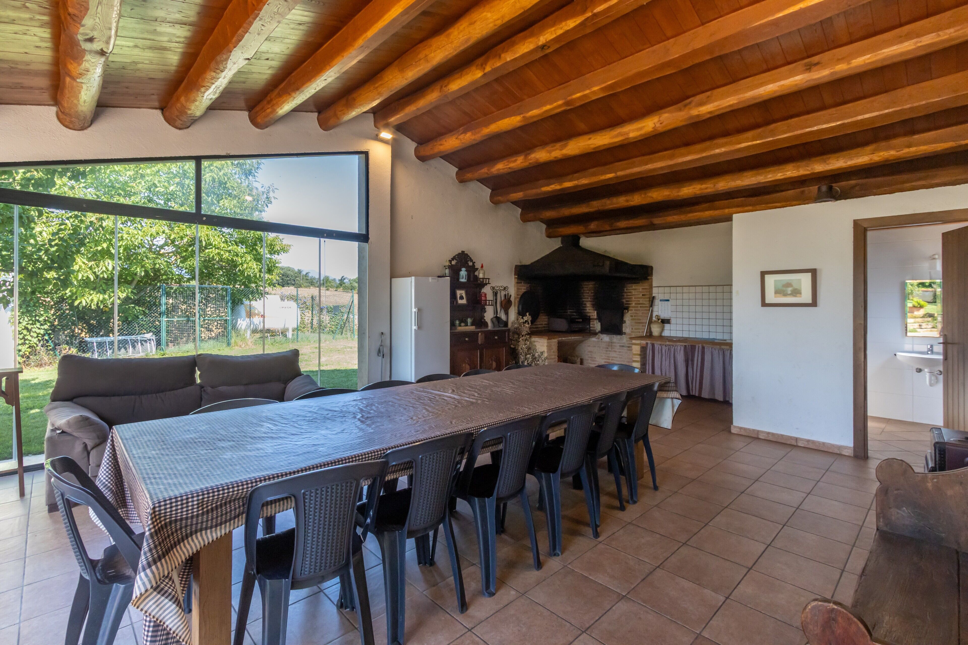 A dining area with a long table, chairs, a wooden ceiling, and a view through a large window.