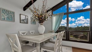 Ein Essbereich mit einem weißen Tisch und Stühlen, einer Vase mit Blumen und einem großen Fenster mit Blick auf Himmel und Wolken.