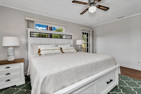 Un dormitorio con una cama, mesitas de noche, ventilador de techo y ventana con vista a vegetación.