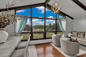 Amplia habitación con un ventanal que ofrece vistas a un campo de golf, una lámpara de araña moderna y una zona de estar acogedora con sofá y sillones.