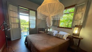 Une chambre à coucher avec un grand lit, un baldaquin, une chaise, une table avec une lampe et une vue sur la verdure à l’extérieur.