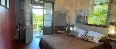 A bedroom with a large bed, a canopy, a window with a view of greenery, and an open door leading to a balcony.