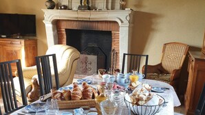 A dining table set with breakfast items, including croissants, bread, and beverages, in a room with a fireplace and wicker chairs.