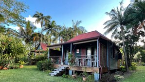 A small, single-story house with a red roof, surrounded by lush greenery and palm trees.