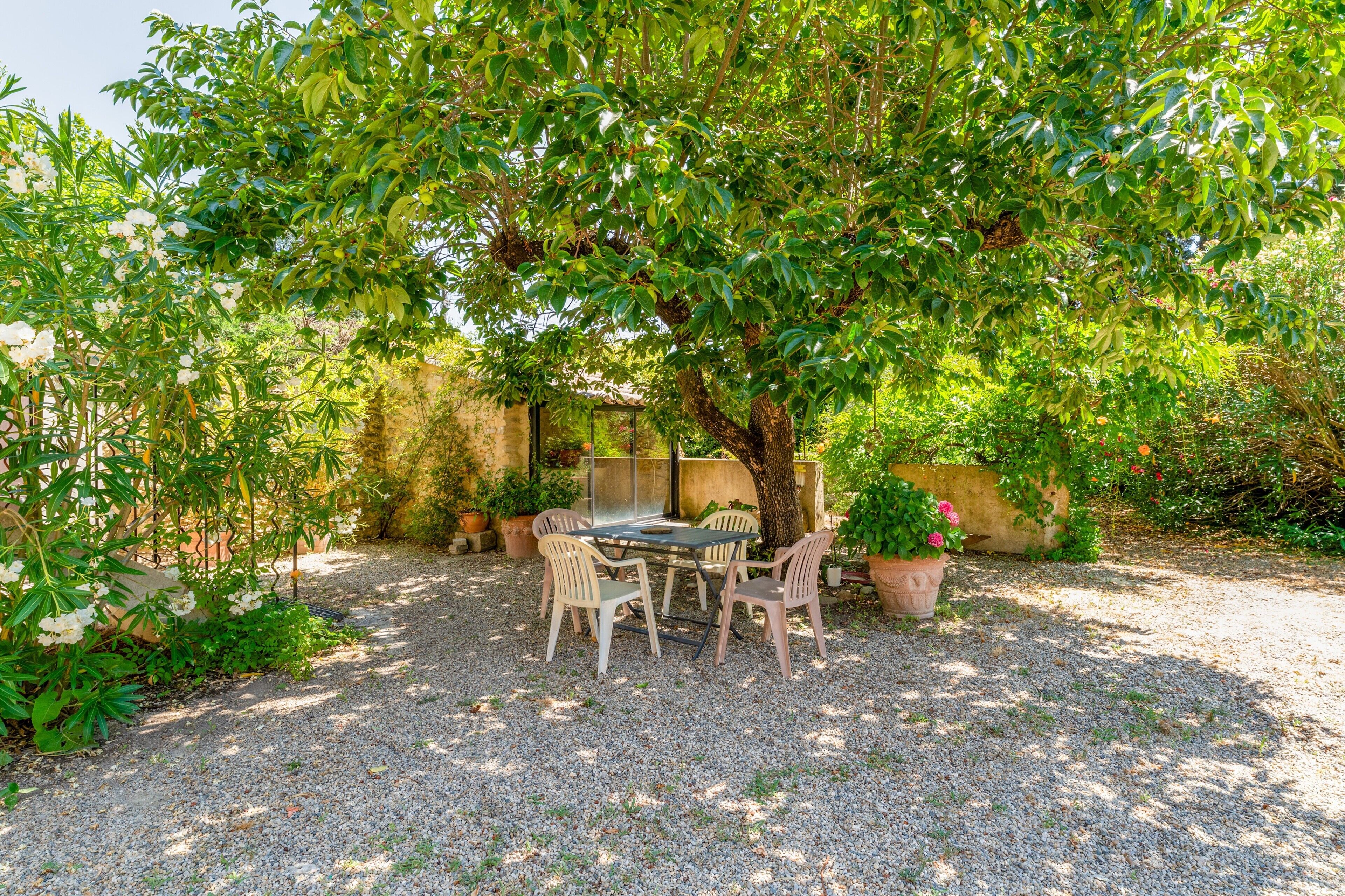 Une terrasse avec une table et des chaises, située sous un arbre, et entourée de gravier et de plantes en pot.