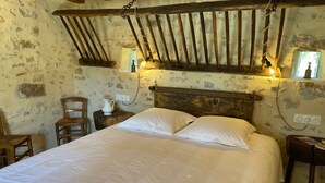 A rustic bedroom with a wooden headboard, a bed with white linens, a small table with a pitcher, and a chair.