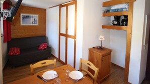 A compact living space with a sofa, a dining table with chairs, a small cabinet, and a wall-mounted shelf. - 2-Room Apartment Close to Slopes (Chamrousse)