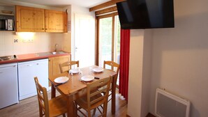 A compact kitchen with wooden cabinets, a microwave, and a built-in oven. A dining table with chairs is set up near a window with red curtains. - 2-Room Apartment Close to Slopes (Chamrousse)