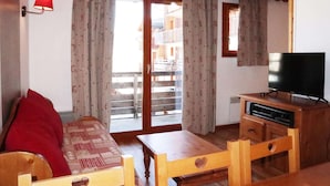 Una habitación acogedora con un sofá de madera, un televisor sobre un mueble de madera y un balcón con cortinas.