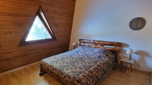Un dormitorio con techo de madera, un tragaluz triangular, una cama con colcha estampada, dos lámparas de noche y un espejo redondo pequeño en la pared.
