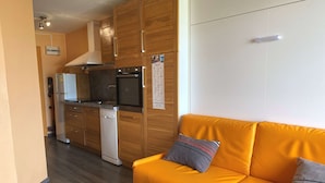 A compact kitchen with wooden cabinets, a built-in oven, and a dishwasher. There is a yellow sofa with two pillows in the living area.