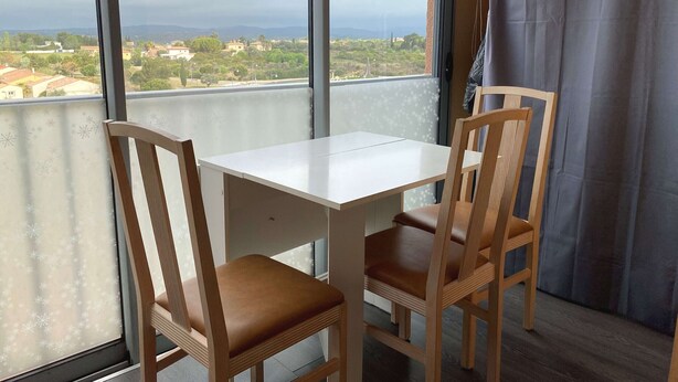 A balcony with a white table and two wooden chairs, overlooking a residential area with houses and trees.