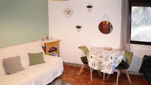 Una sala de estar acogedora con un sofá blanco, una mesita con sillas, un espejo redondo y un ventanal con vistas a árboles.