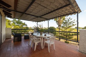 Une terrasse extérieure avec une table ronde et des chaises blanches, entourée d’une rambarde en métal et offrant une vue sur la verdure.
