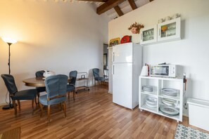 A dining area with a round table, chairs, a refrigerator, a microwave, and a shelf with dishes.