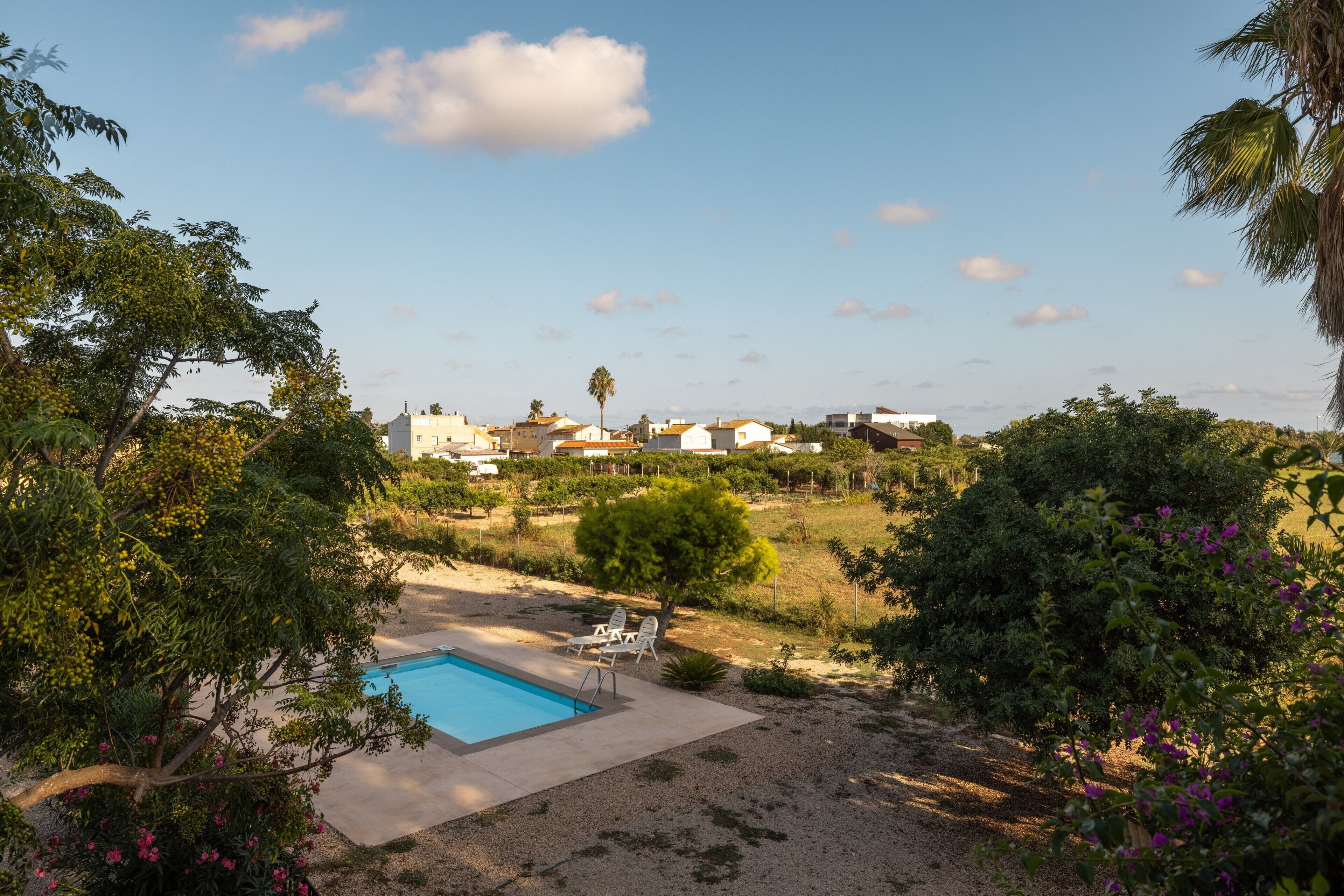 A swimming pool with lounge chairs, surrounded by greenery and buildings in the background.