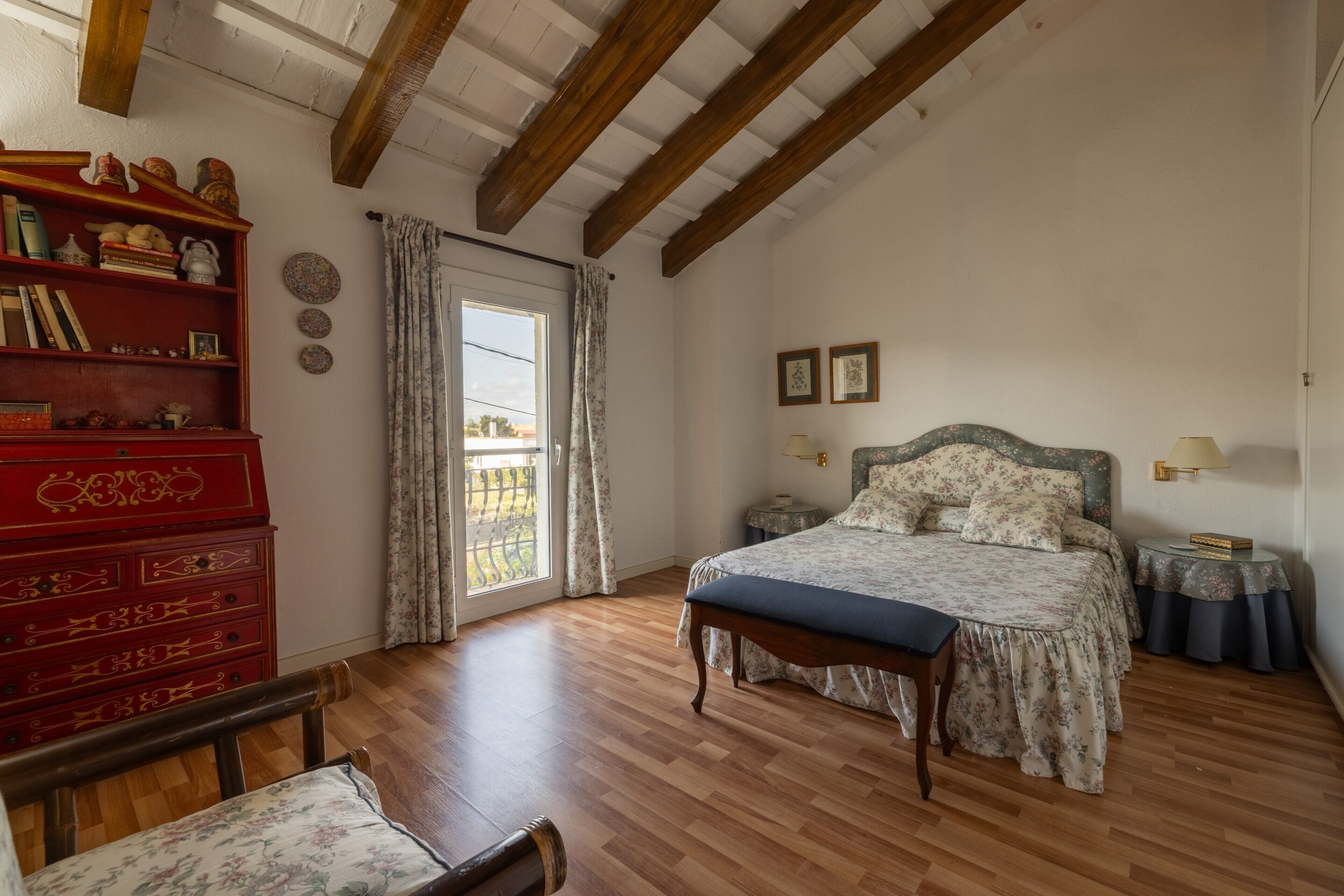 Un dormitorio con suelo de madera, una cama con ropa de cama floral, un tocador rojo y una ventana con cortinas.
