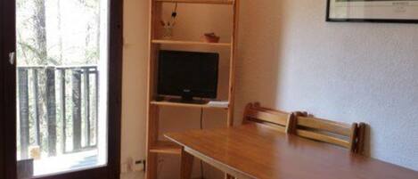 Une pièce avec une table et des chaises en bois, un téléviseur posé sur une étagère et une fenêtre donnant sur des arbres.