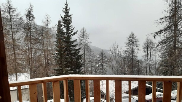 A snowy outdoor scene with a wooden railing, a view of parked cars, and a forested area with trees.