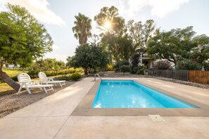 Une piscine rectangulaire avec un escalier en métal, entourée d’une terrasse en béton. On aperçoit des chaises longues et une clôture en bois à l’arrière-plan.