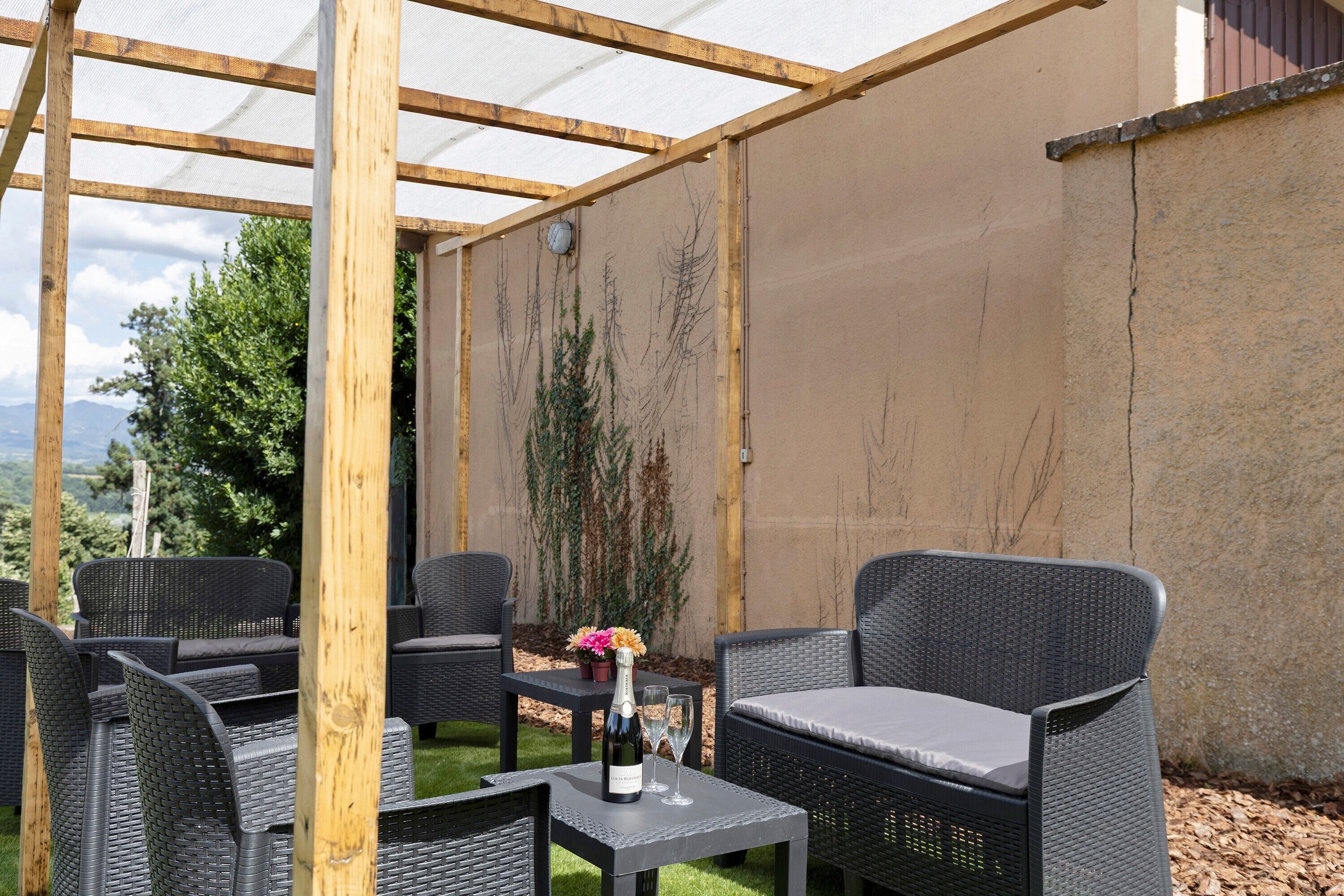 Un espacio de estar al aire libre cubierto, con mobiliario de mimbre y una mesa con una botella y vasos.