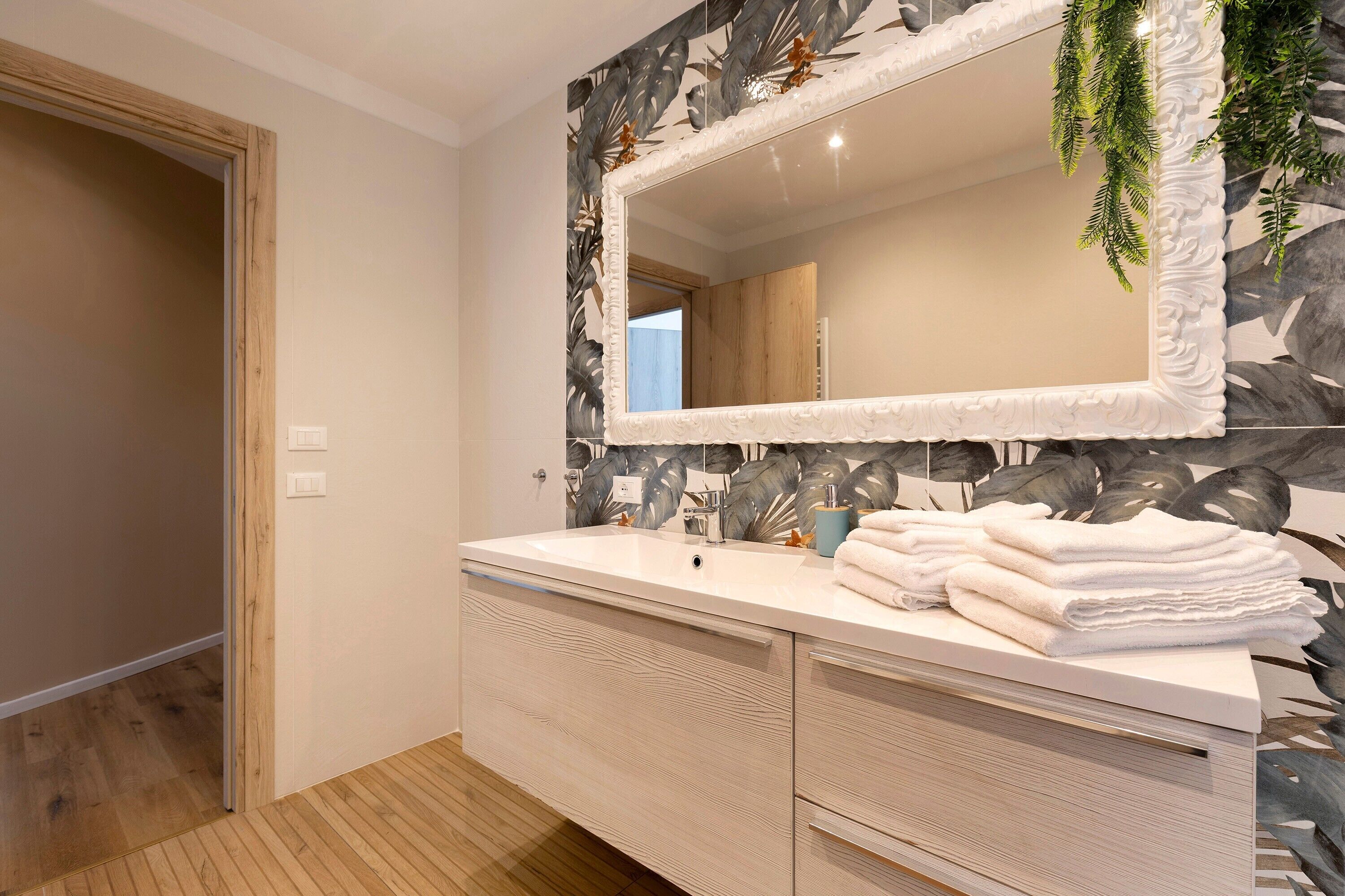 A bathroom with a wooden floor, a white vanity with a mirror, and a wall with a leaf pattern.