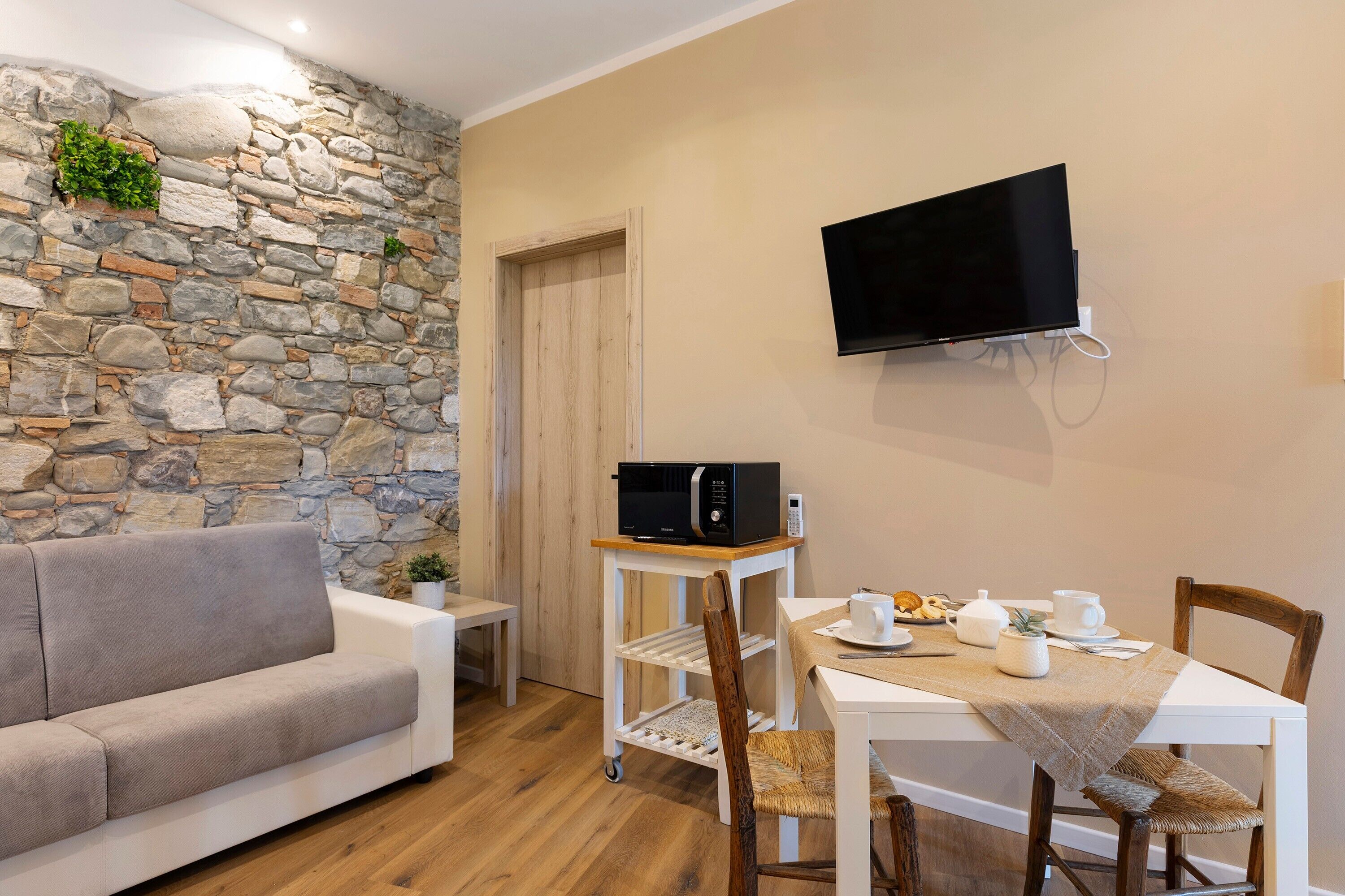 A cozy living area with a stone wall, a flat-screen TV, a white table with chairs, a sofa, and a small cart with a microwave.