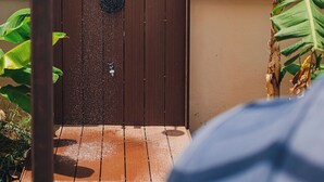 Une douche extérieure en bois, dotée d’un jet d’eau vertical, entourée de plantes et agrémentée d’une terrasse en bois.