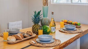 Un coin repas avec une table en bois, deux lampes orange suspendues, un vase contenant des plantes séchées, un ananas, une bouteille de Sol y Sol et une fenêtre donnant sur un paysage.