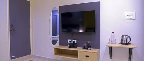 Une chambre d’hôtel moderne équipée d’un téléviseur à écran plat fixé au mur, d’une console en bois sur laquelle sont posés une bouilloire et des verres, et d’un petit miroir également fixé au mur.