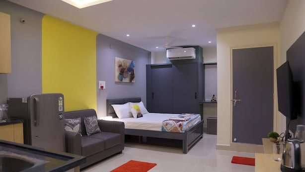 Quarto de hotel moderno com cama, sofá, pequena cozinha e televisão.