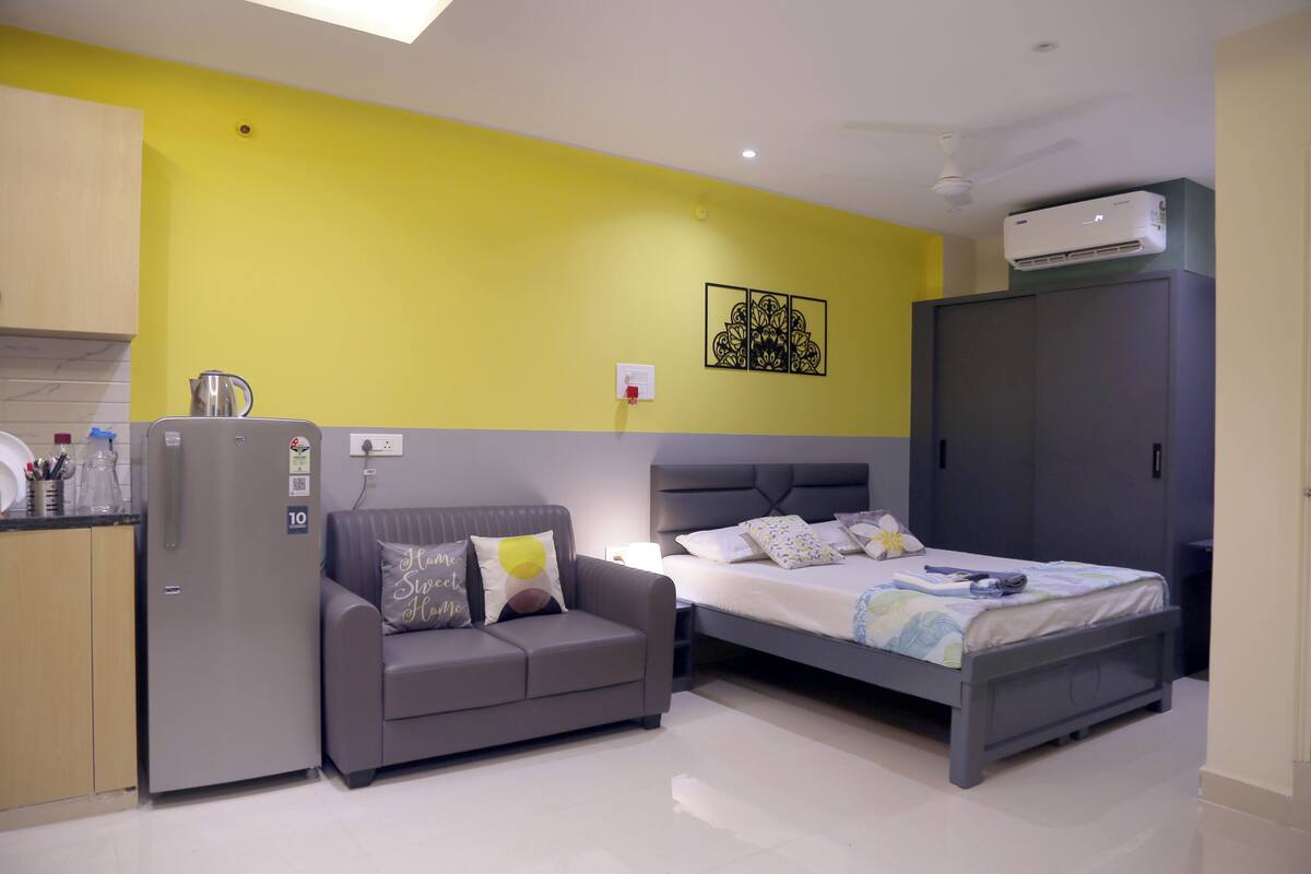 A compact hotel room with a kitchenette, a sofa, a bed, and a wardrobe.
