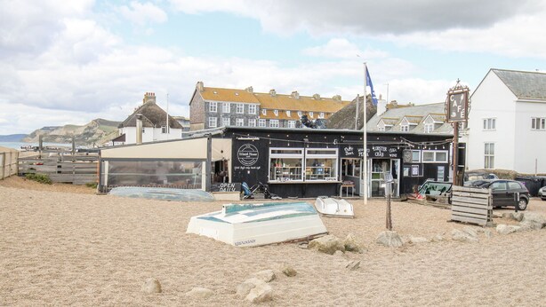 A coastal building with a signboard advertising a cafe, a visible boat, and a rocky beach. - Beachcombers, 13 Chesil House (Bridport)