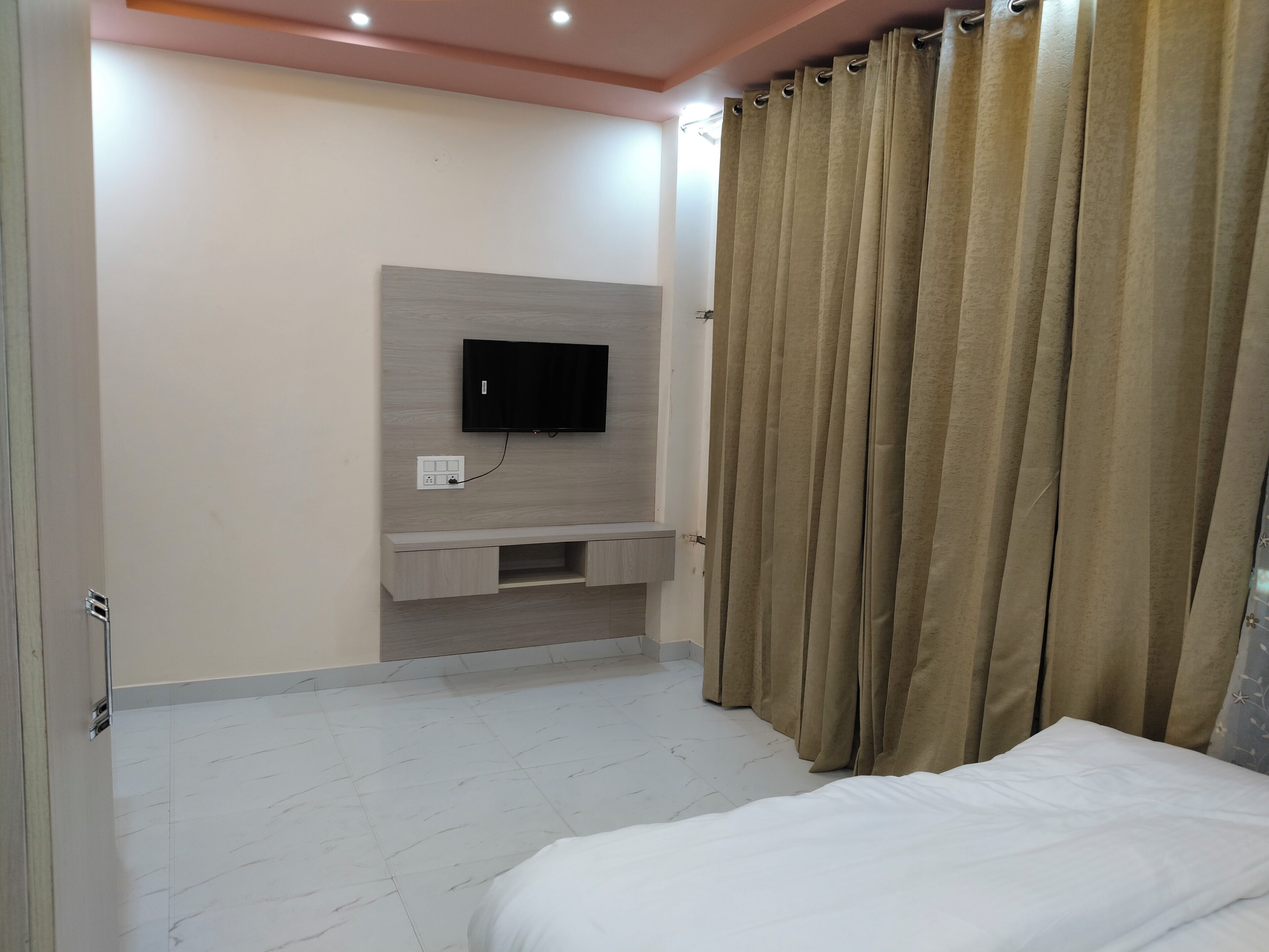 A bedroom with a bed, a TV mounted on the wall, a small cabinet, and a curtain.