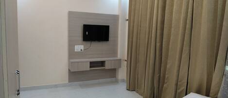 A bedroom with a bed, a TV mounted on the wall, a small cabinet, and a curtain.