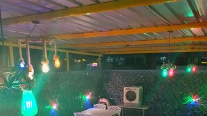 A restaurant with white metal tables and chairs, hanging light fixtures, and a visible air conditioning unit. - Modest Rooms by LIBRA (Kharar)