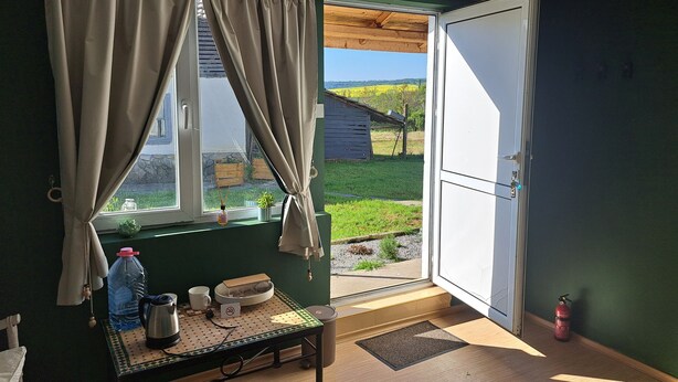 A room with a table, a window with curtains, a door, and a view of a green field and a wooden structure outside. - Muddy Puddles Farm (Kovachevets)