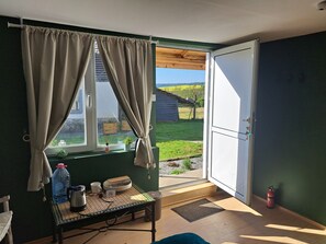 A room with a table, a window with curtains, a door, and a view of a green field and a wooden structure outside. - Muddy Puddles Farm (Kovachevets)