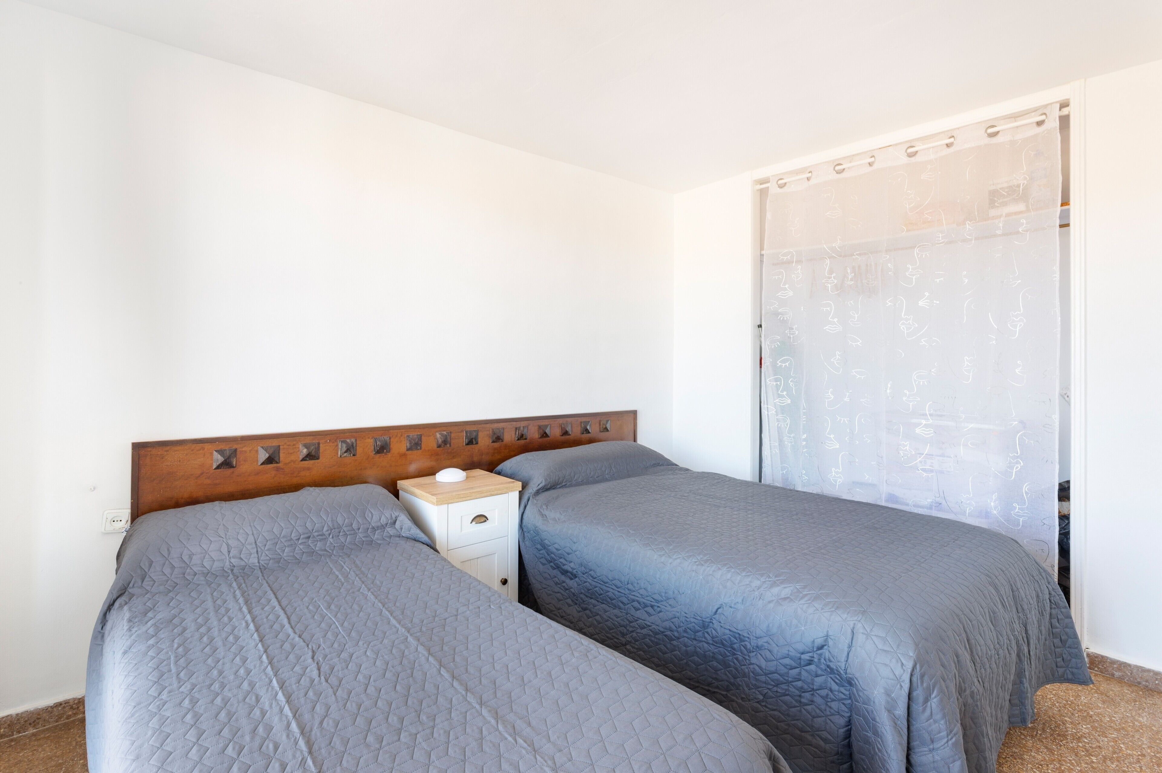 A bedroom with two beds, a wooden headboard, a bedside table, and a curtain with a pattern.