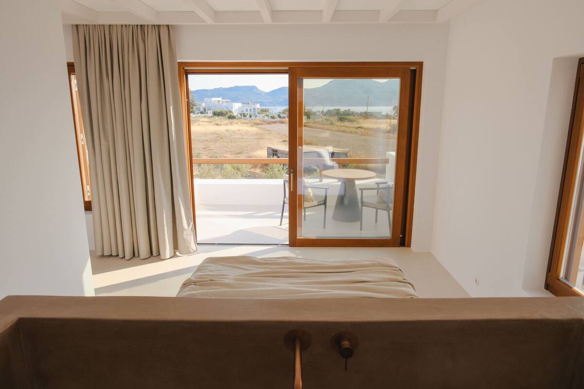 A bedroom with a large window, a bed, and a view of a balcony with outdoor furniture.