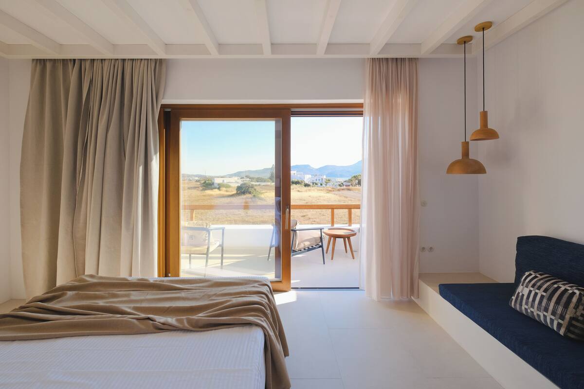 A bedroom with a bed, a sofa, a balcony with a table and chairs, and a view of the landscape.